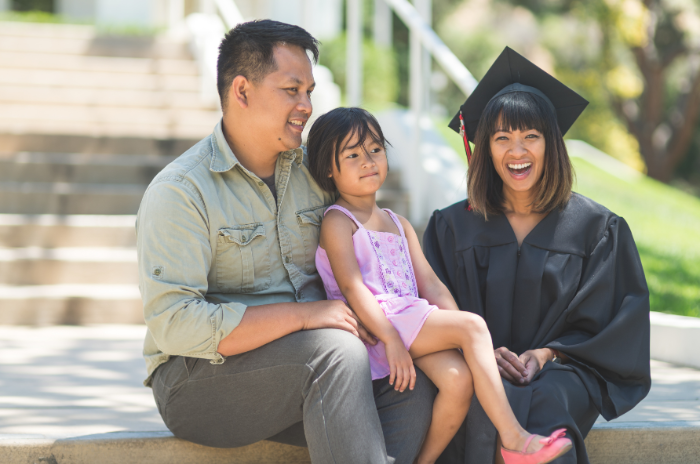 A woman sits on steps after graduation with her husband and their young daughter. Her daughter is sitting on dad's lap.
