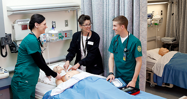 teacher and students standing over a dummy lying in a hospital bed