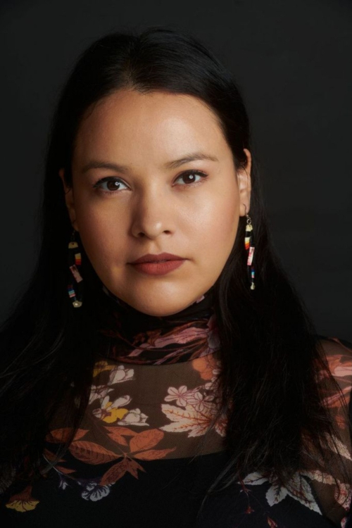 A young, dark-haired woman, head slightly tilted, wearing a high floral top and beaded earrings.