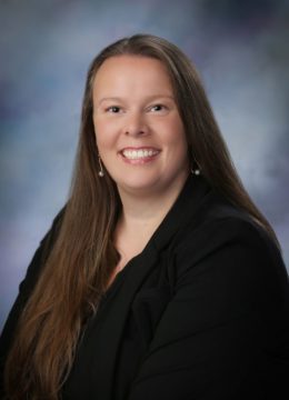 A smiling women with dark hair and a deep blue shirt, set in front of a studio backdrop.
