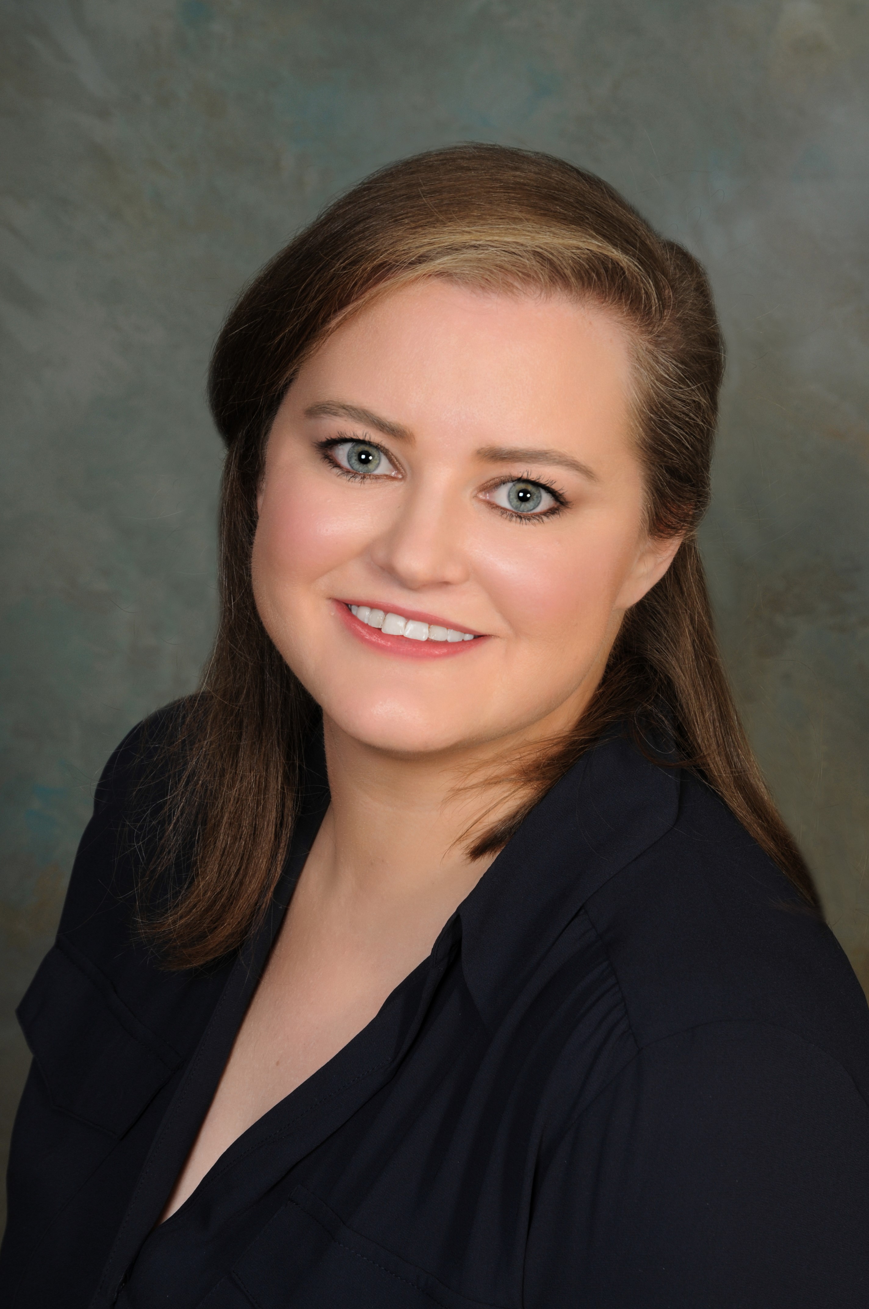 An angled view of smiling woman with light brown hair and a dark shirt, standing against a studio backdrop.
