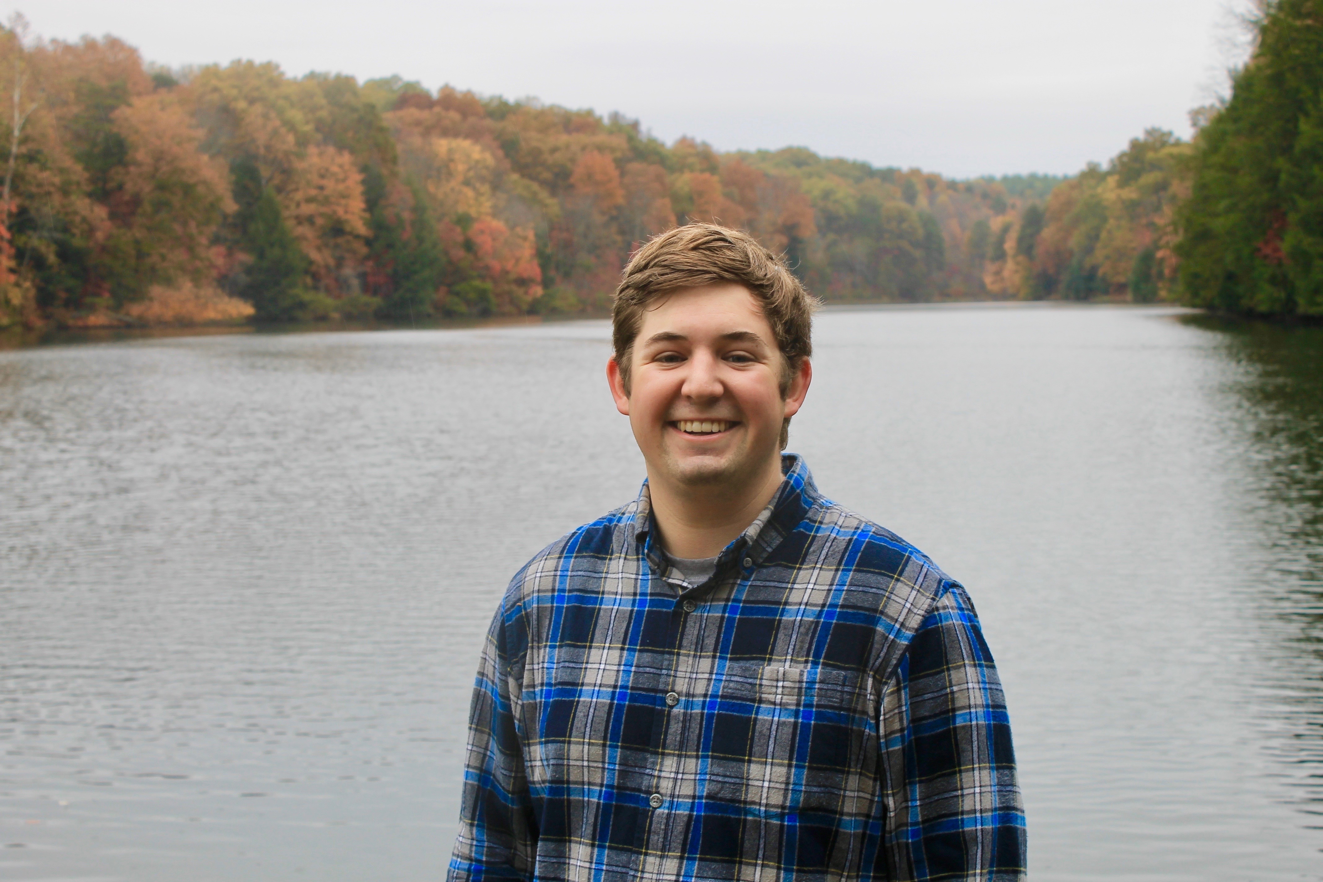 A young, smiling man with thick chestnut hair and a flannel shirt, with a river and autumn trees behind him. 