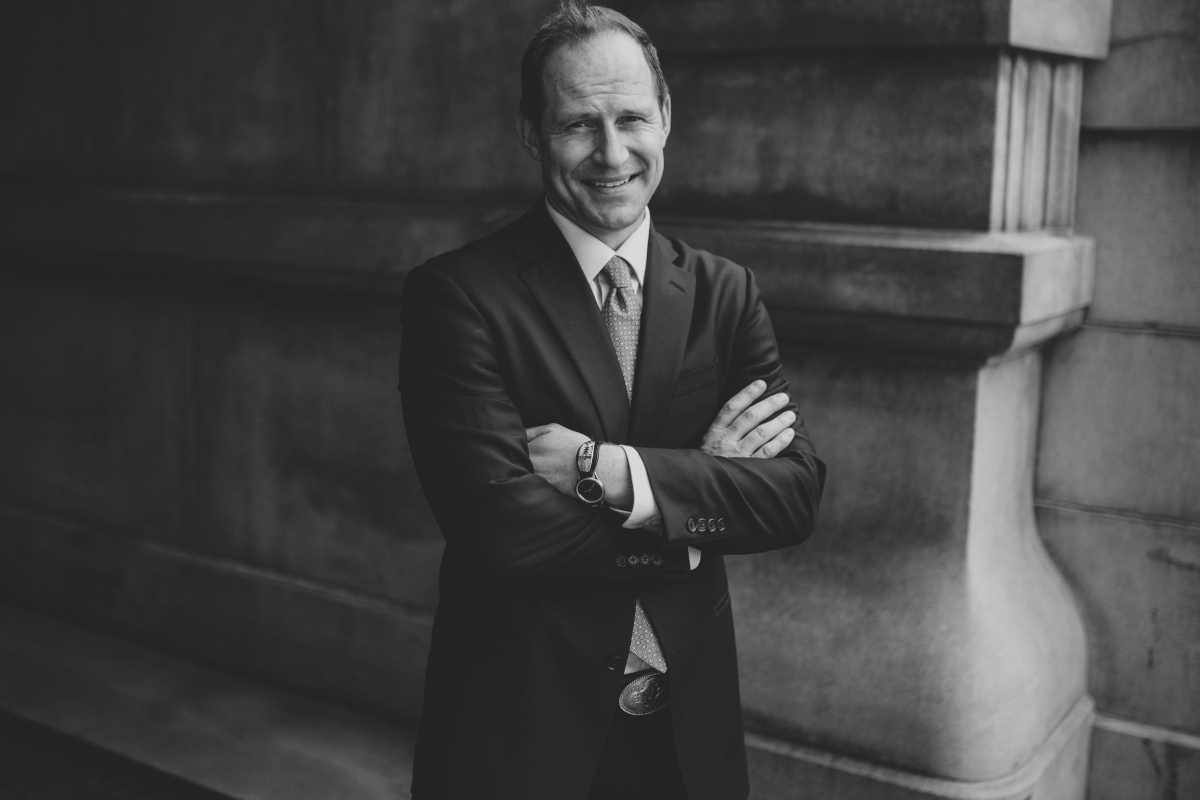 A smiling man, hair tussled and arms crossed, wearing a suit and belt buckle.