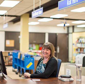 A librarian smiling at the camera from behind the library service desk.